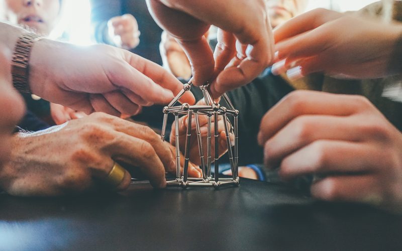 Molte mani attorno a una piccola torre di aste metalliche che stanno montando insieme