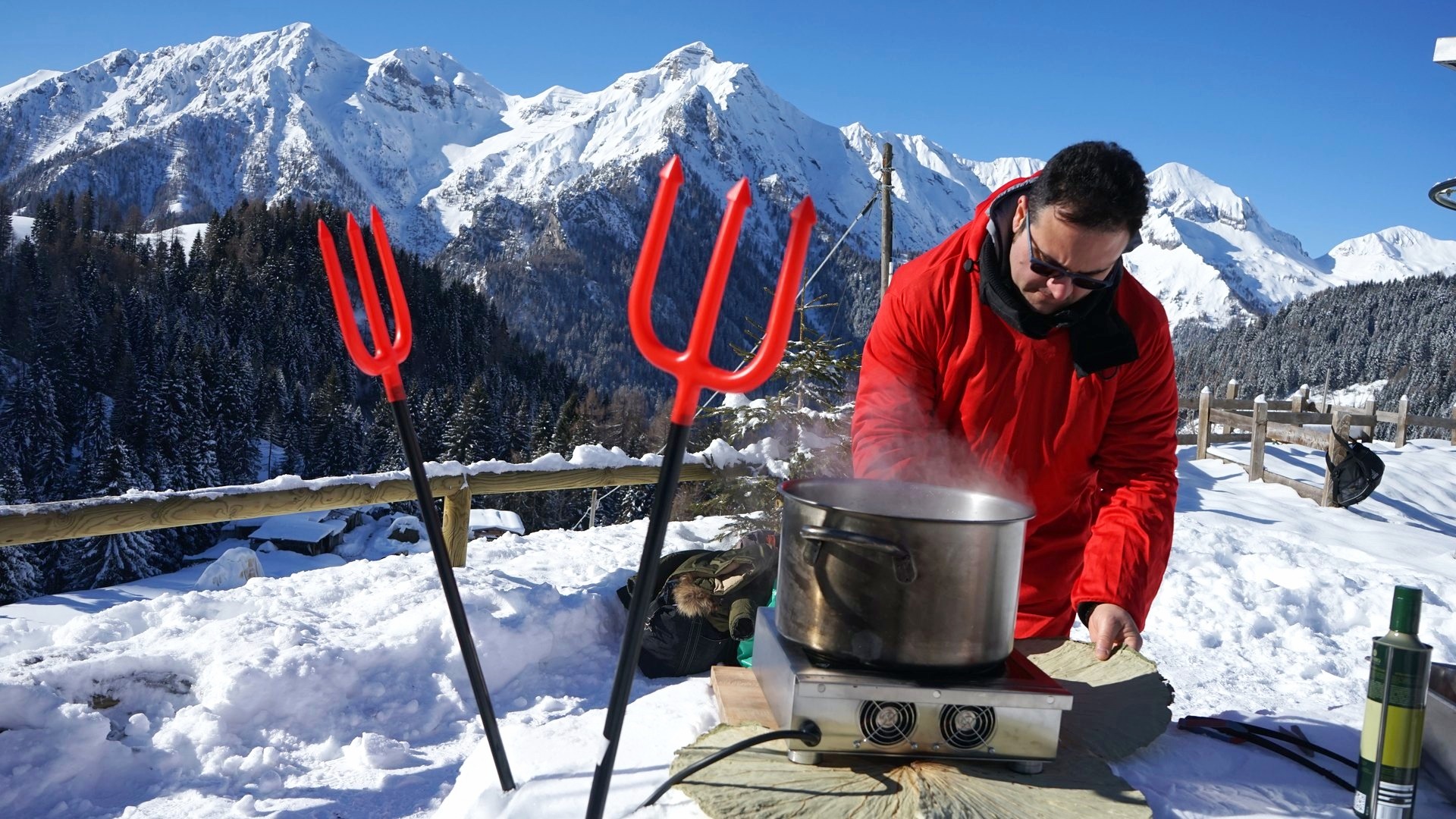 Un cuoco in giacca rossa lavora alla pentola fumante su un terrazzo innevato, con le cime delle Alpi dietro
