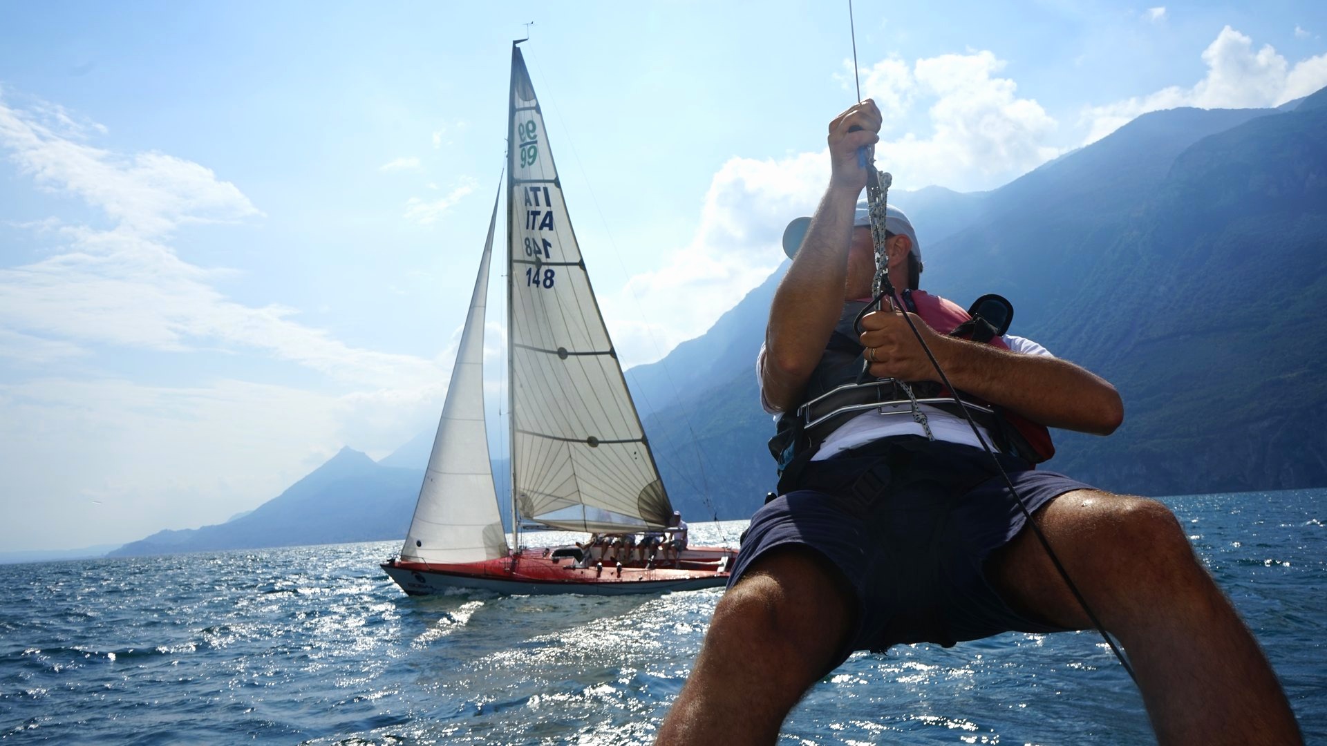 Un membro dell'equipaggio recupera una scotta a bordo, con la barca sotto vela sul lago