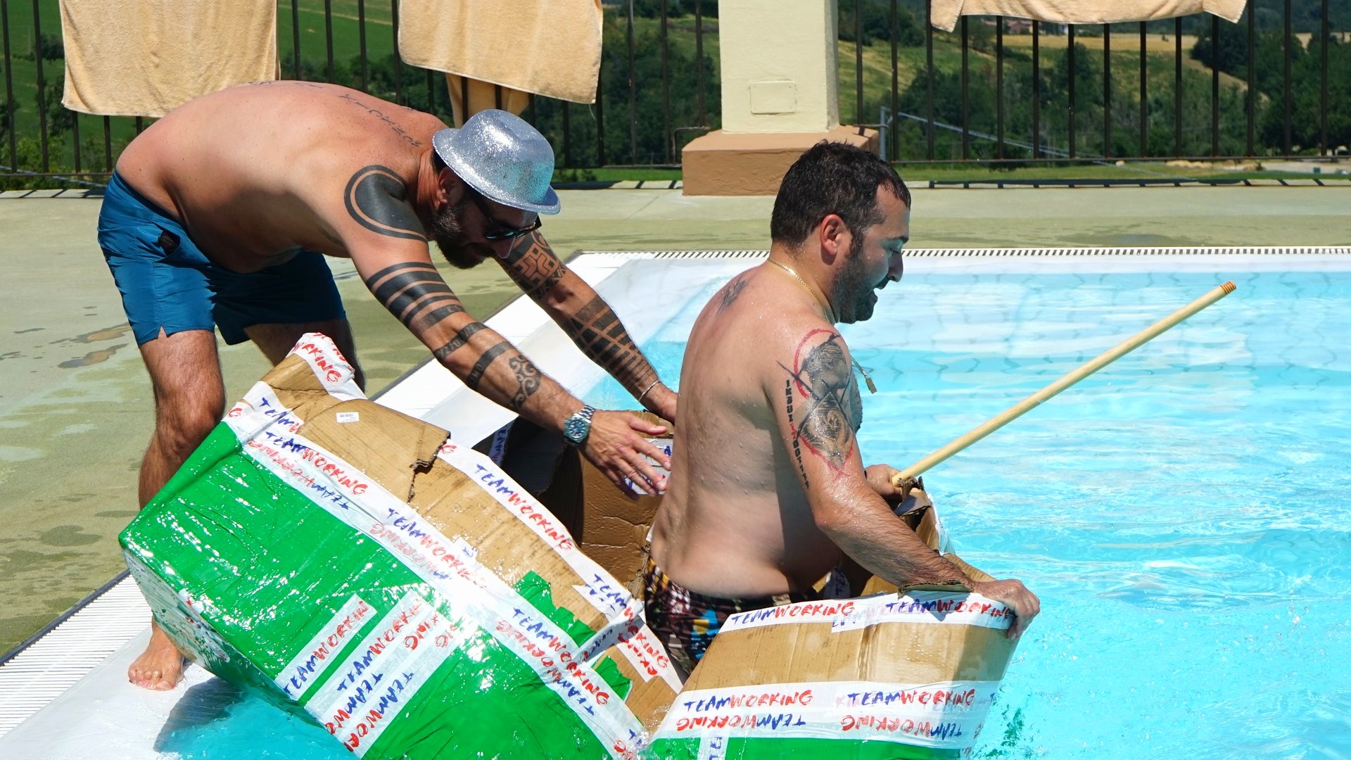 Due partecipanti navigano in piscina su una barca costruita con cartone e nastro adesivo
