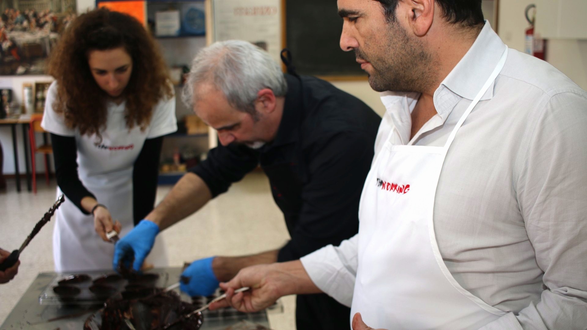Tre partecipanti in grembiule lavorano il cioccolato al banco di un laboratorio