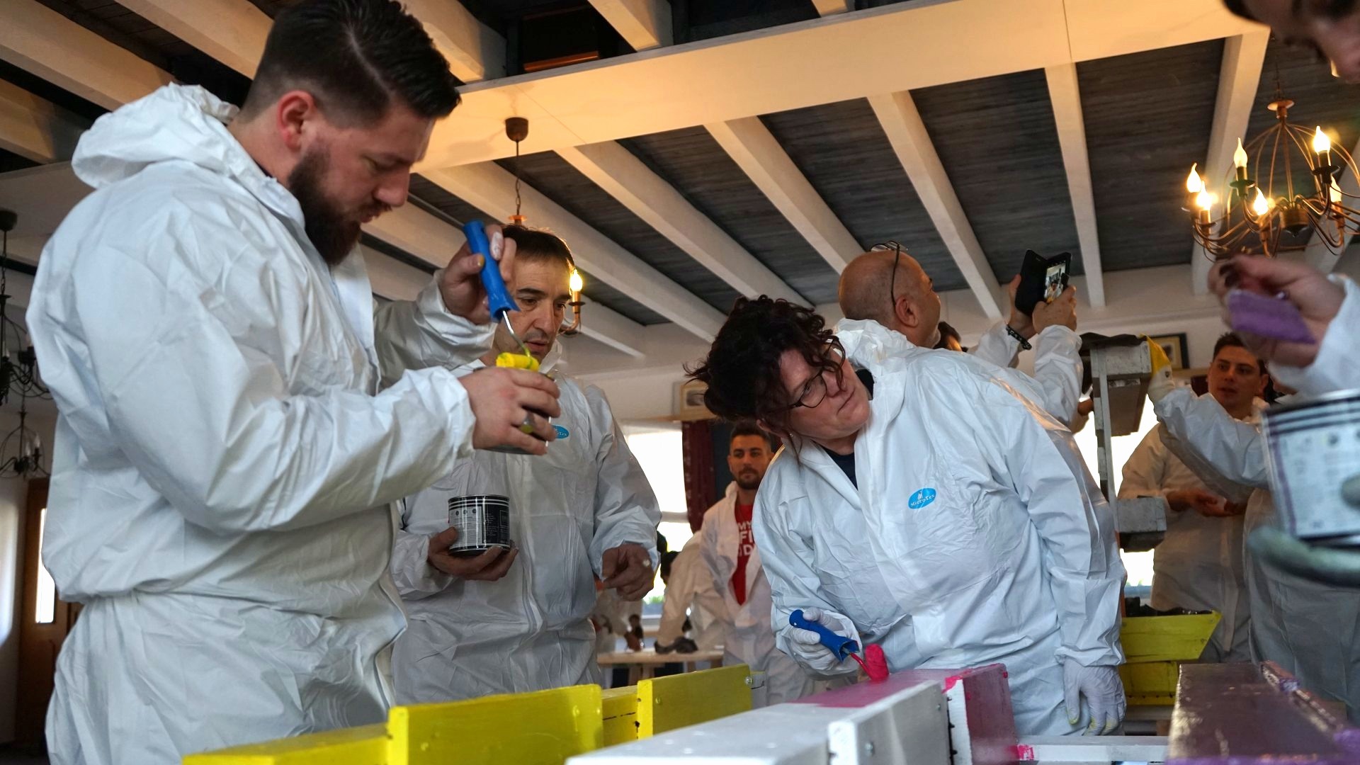 Partecipanti in tuta da lavoro costruiscono e dipingono arredi per un progetto sociale